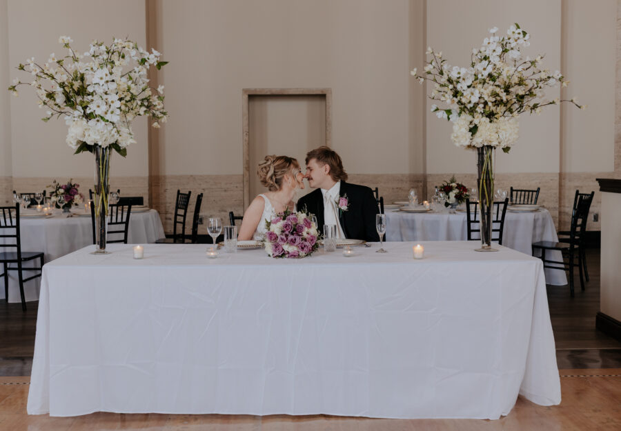 Newlywed couple leaning in for a kiss at their sweetheart table surrounded by tall floral arrangements.